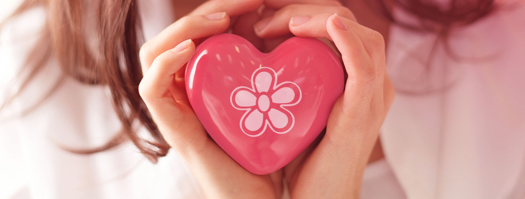 Dettaglio sulle mani di una donna che stringe un cuore di marmo dipinto di rosa scuro e con impresso al centro in bianco il simbolo della margherita del logo di APE